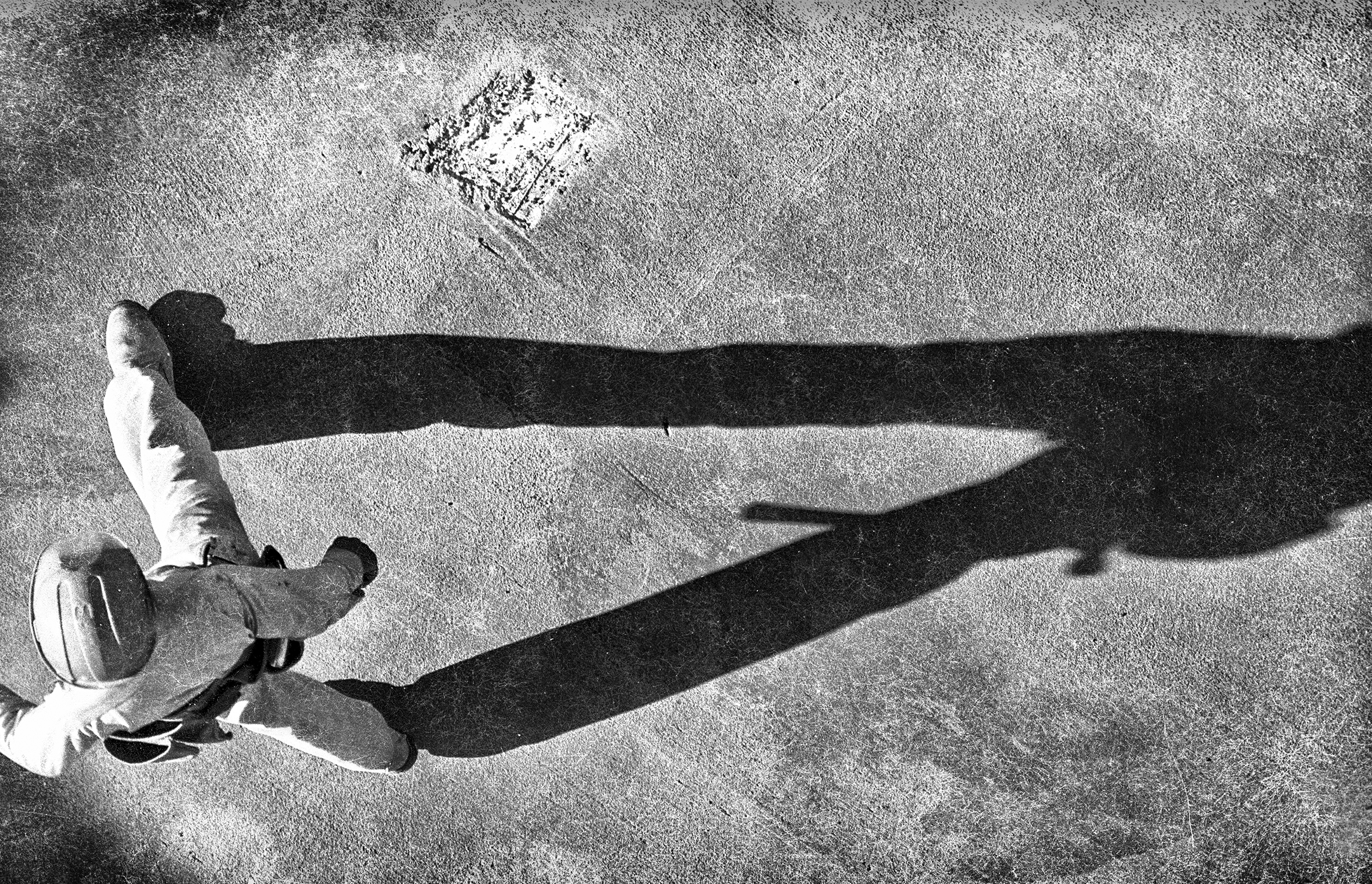 A workman in a hard hat wearing a tool belt is seen from above walking on a concrete floor as his shadow looms large to the right.
