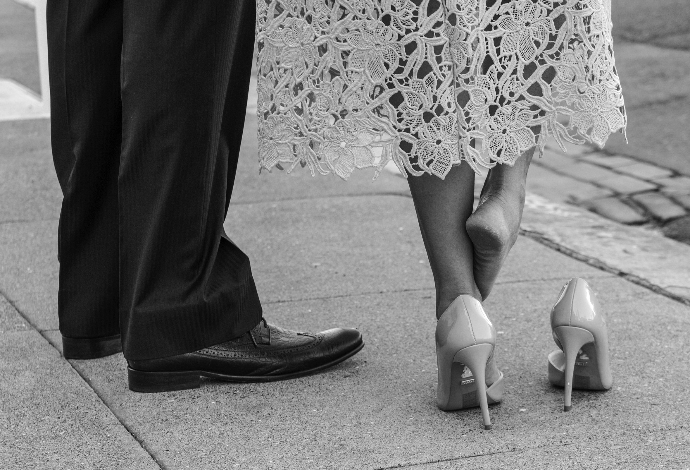 A man and woman stand on a sidewalk. Seen from the knees down, he is wearing dress pants and shoes, she is wearing a midcalf linen dress with flowered lace above the hem and high heels. Her right foot is out of its shoe and is rubbing her left ankle.