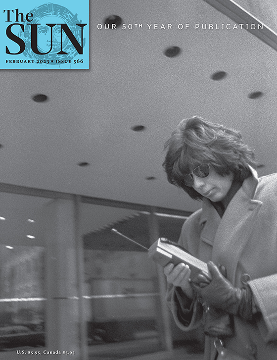 February 2023 cover of The Sun. A woman in Manhattan in the 1990s stands in front of a building looking down at the very large mobile phone she is holding. She is wearing a winter coat and has removed the glove from her right hand.